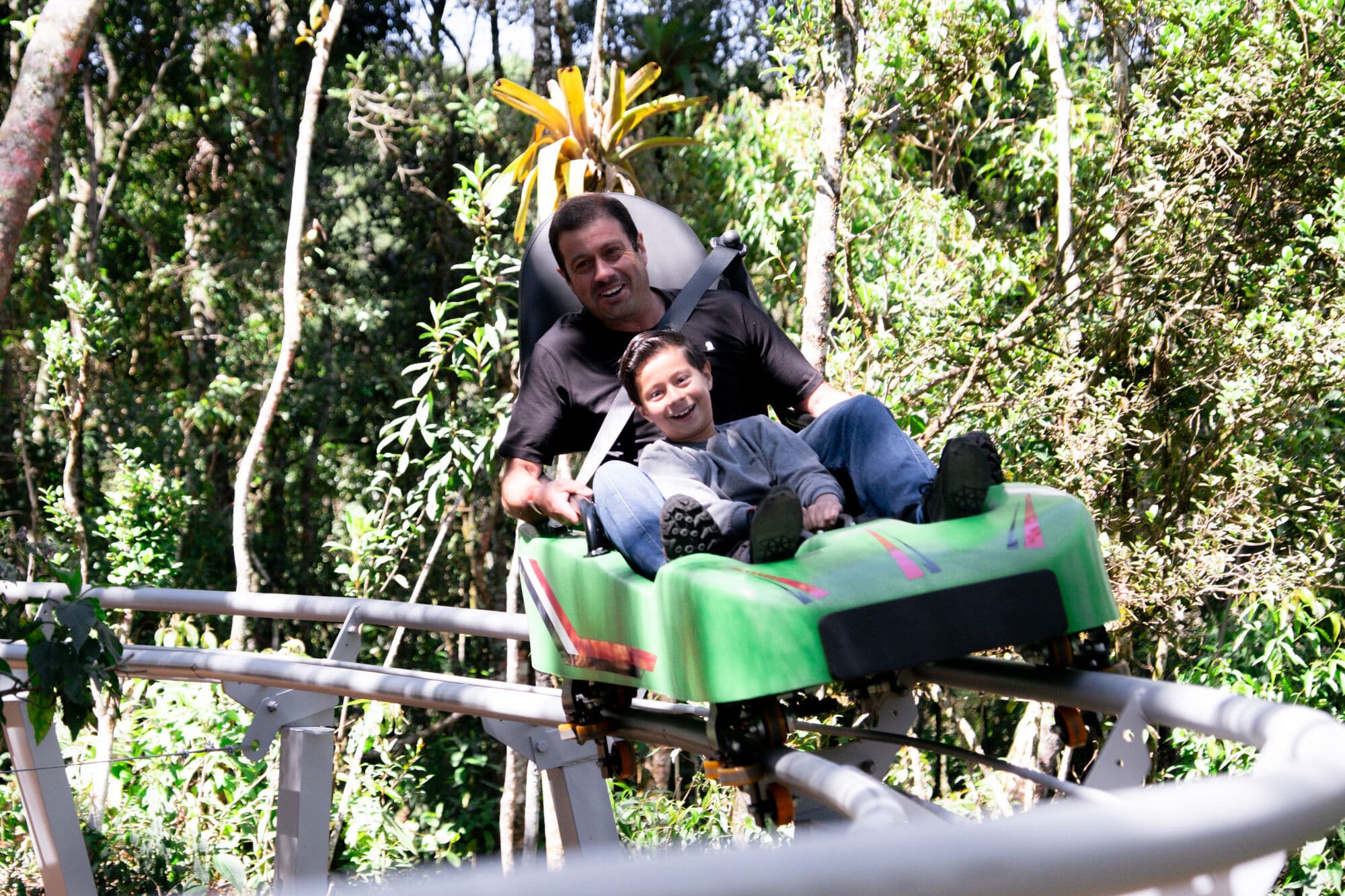 Pai e filho sorriem descendo juntos no trenó de montanha em meio à mata